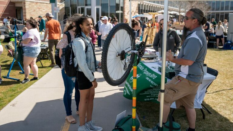 Student stands in front of a bike tire suspended on a pole as a worker talks to them. They stand in front of the LSC Sutherland Gardens, where many people are walking and interacting in the background.